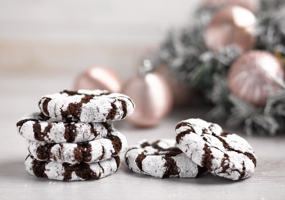 Baker & Baker Snow Topped Crinkle Cookies a pile of six brown and white festive cookies infront of a frosted white christmas tree decorated with gold baubles