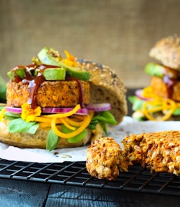 Paramount’s BBQ Lentil & Mixed Seed Burger With Chargrilled Avocado & Caramelised Onion Hummus two burger buns filled with lentil and mixed seeds burgers, avocado, sauce and salad on a wire cooling rack
