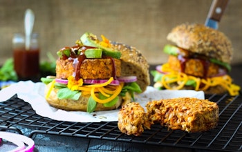 Paramount’s BBQ Lentil & Mixed Seed Burger With Chargrilled Avocado & Caramelised Onion Hummus two burger buns filled with lentil and mixed seeds burgers, avocado, sauce and salad on a wire cooling rack