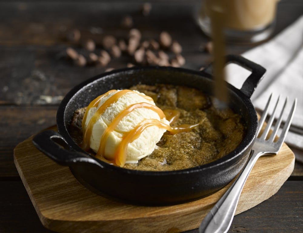 black skillet with warm cookie dough with a scoop of vanilla ice cream and chocolate sauce drizzled on top 