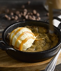 Baker & Baker Warm Cookie Dough Dessert black skillet with warm cookie dough with a scoop of vanilla ice cream and chocolate sauce drizzled on top