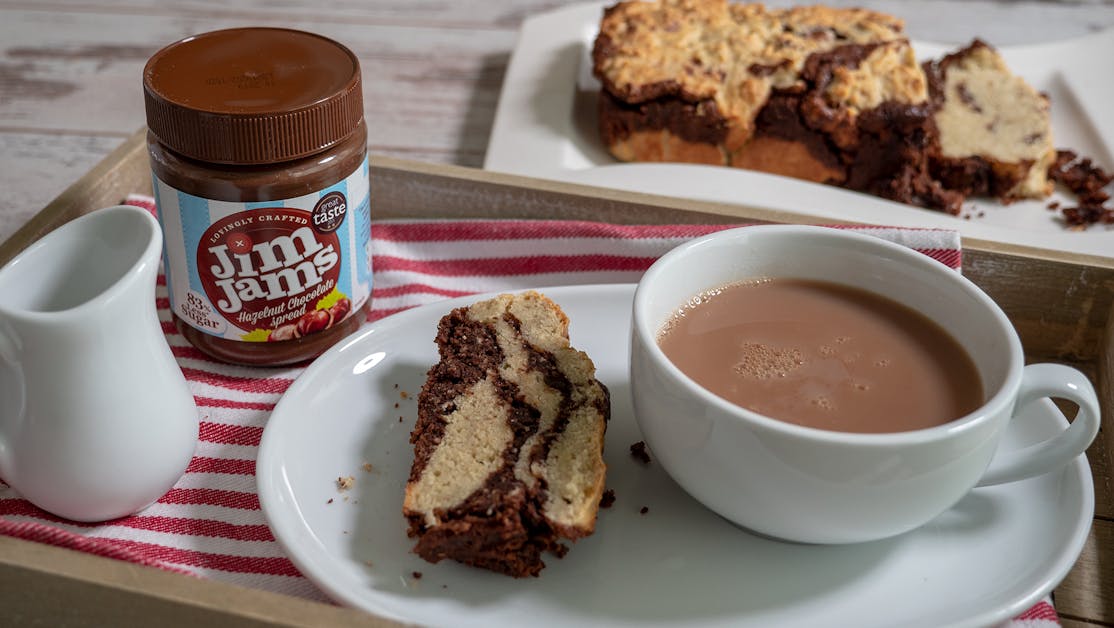 JimJams Chocy Banana Bread a chocolate banana bread loaf placed behind a dining tray with a jar of jimjams chocolate spread, a slice of the cake, and a cup of tea