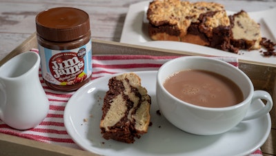 a chocolate banana bread loaf placed behind a dining tray with a jar of jimjams chocolate spread, a slice of the cake, and a cup of tea 