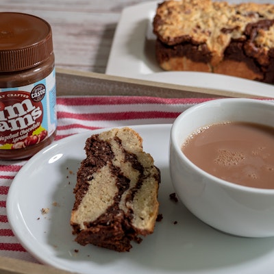 a chocolate banana bread loaf placed behind a dining tray with a jar of jimjams chocolate spread, a slice of the cake, and a cup of tea 