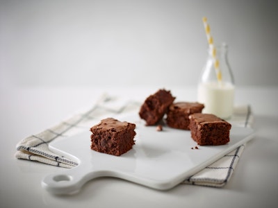 chunks of brownie on a glossy white chopping board next to a glass of milk with a yellow and white straw 
