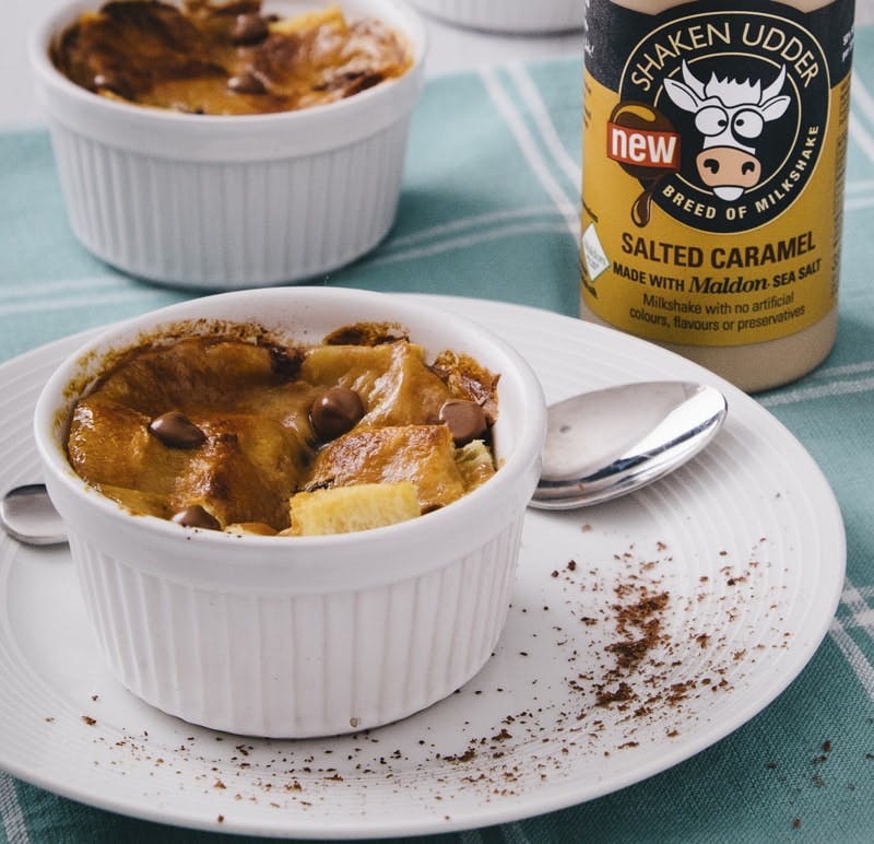Salted Caramel Bread & Butter Pudding white ramekin on a white plate filled with salted caramel bread and butter pudding next to a bottle of shaken udder salted caramel milkshake