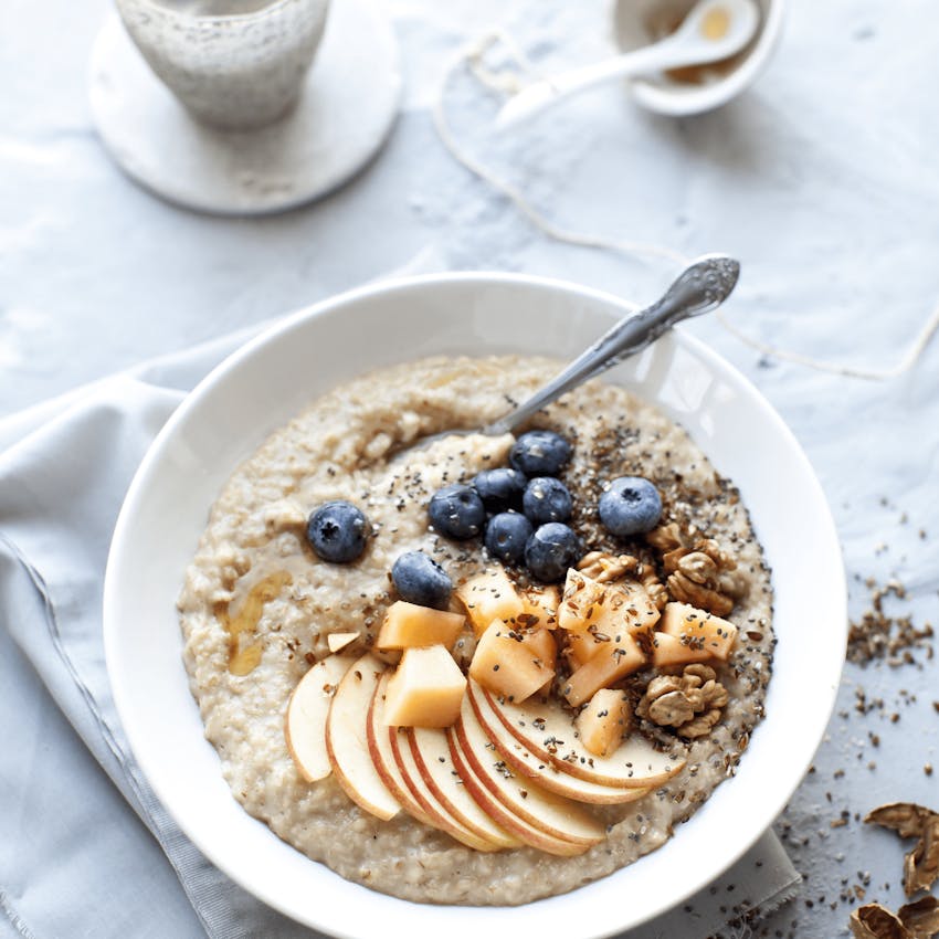 a white bowl filled with creamy porridge topped with stewed apples, blueberries nuts and seeds