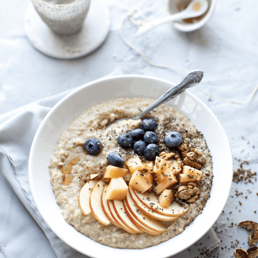 Delicious, fruit topped porridge. a white bowl filled with creamy porridge topped with stewed apples, blueberries nuts and seeds