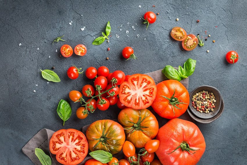 birds eye view of cherry vine and beefsteak tomatoes scattered across a grey marble tabletop and a pot of tomato seeds