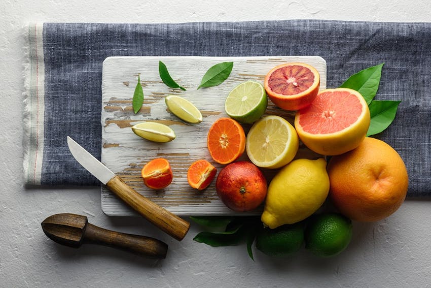 light wooden chopping board on top of a table runner topped with whole and halved limes, lemons and orange citrus fruits 