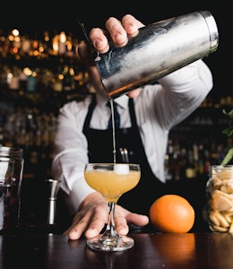 bartender holds a cocktail shaker over a half serve glass pouring in an alcoholic drink