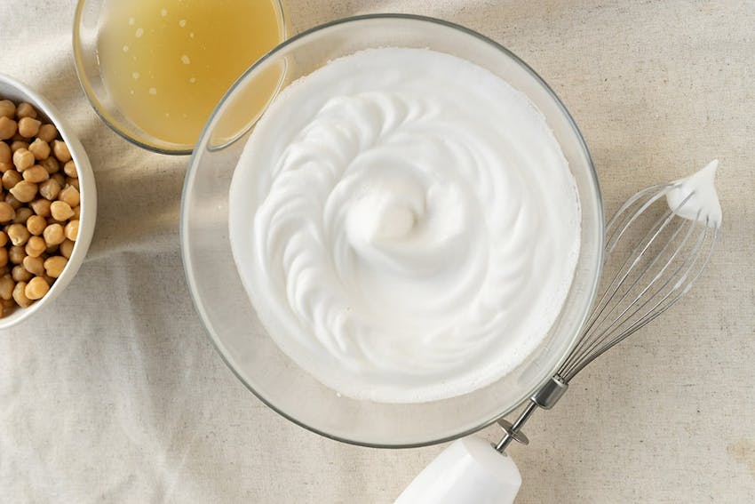 clear bowl of fluffy creamy whisked chickpea water next to a whisk a bowl of chickpeas and chickpea water 