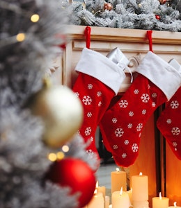 three red christmas stockings hung on the fireplace above candles next to a frosted christmas tree with red and gold baubles 