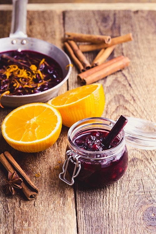 a jar of cranberry sauce on a wooden table next to a sliced orange and cinnamon sticks 