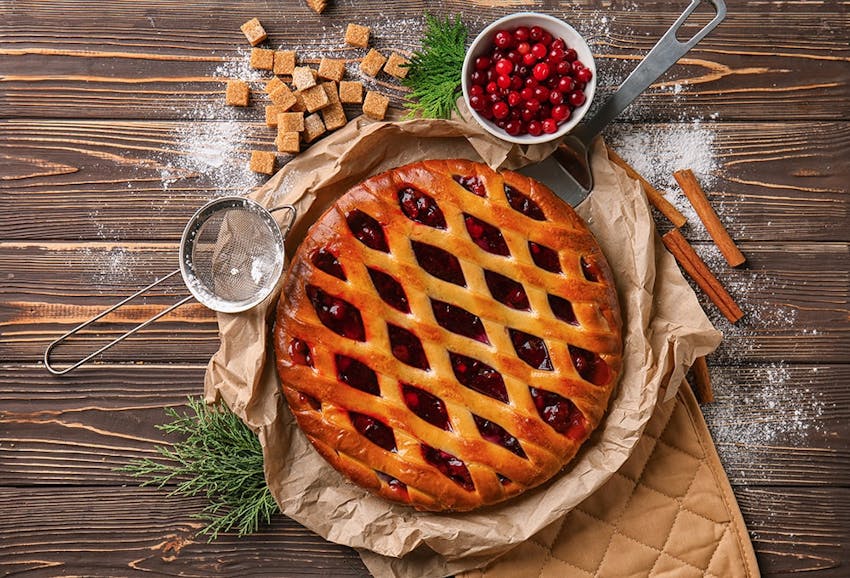 crispy golden christmas pie with a latticed top and a layer of cranberry sauce placed on a dish with brown baking paper on top of an oven glove 