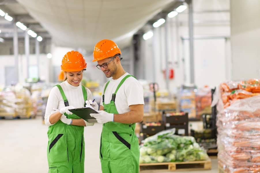 a man and women in white tops green dungarees and orange hard hats look at a clipboard in the middle of a food wholesaler warehouse 