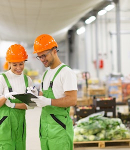 Wholesaler Update Part 2 a man and women in white tops green dungarees and orange hard hats look at a clipboard in the middle of a food wholesaler warehouse