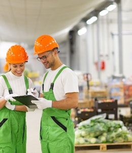 a man and women in white tops green dungarees and orange hard hats look at a clipboard in the middle of a food wholesaler warehouse 