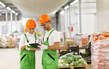 a man and women in white tops green dungarees and orange hard hats look at a clipboard in the middle of a food wholesaler warehouse 