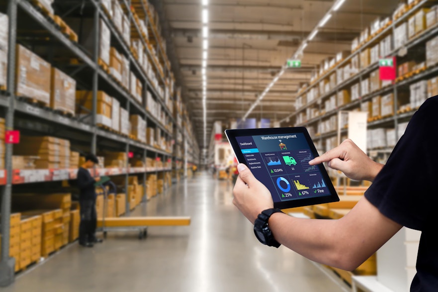a man holds an iPad with a data report infront of stacked shelves inside a food wholesaler warehouse  