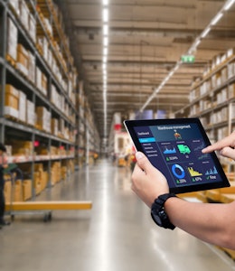 a man holds an iPad with a data report infront of stacked shelves inside a food wholesaler warehouse  