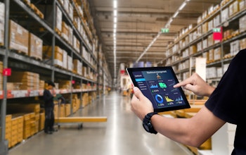 a man holds an iPad with a data report infront of stacked shelves inside a food wholesaler warehouse  