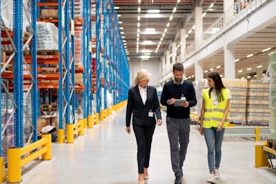 a woman in a suit and a woman in a green high vis walk through a food wholesaler warehouse with a man in a jumper holding an ipad