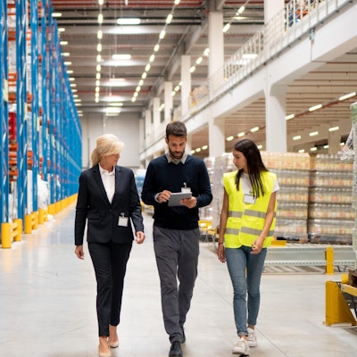 a woman in a suit and a woman in a green high vis walk through a food wholesaler warehouse with a man in a jumper holding an ipad
