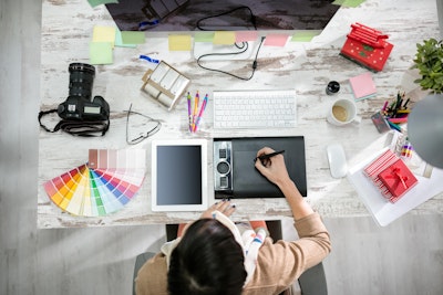 a graphic designer sits at a desk surrounded by design tools, pens, pecils, iPad a camera sketchpad and colour swatches 