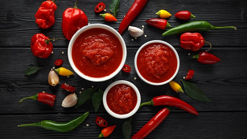 three small white pots of tomato sauce on a black wooden table surrounded by chillis garlic and peppers 