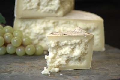 a triangular segment of creamy textured cheese in front of a bunch of green grapes and a cheese wheel