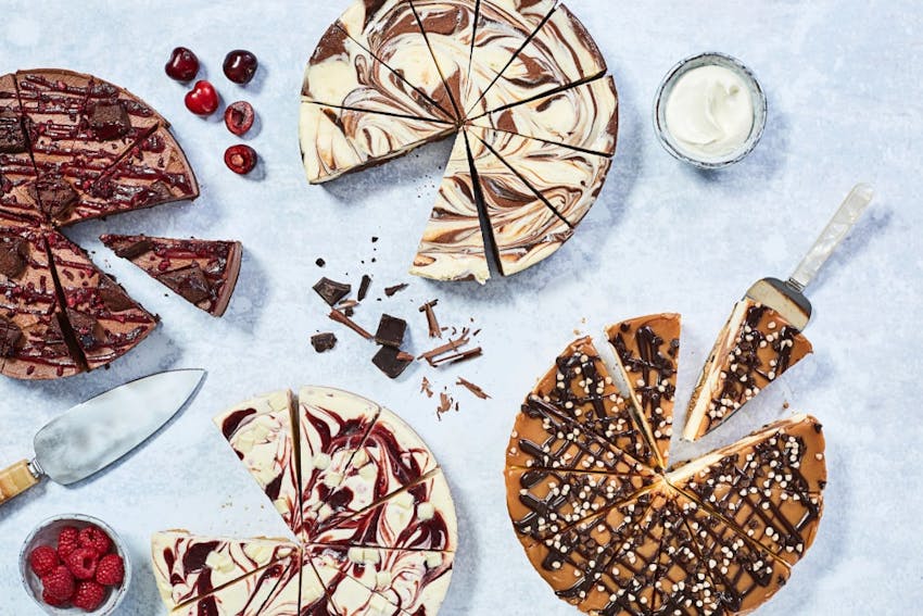 four varying flavours of cheesecakes all equally sliced into 12 slices on a marble tabletop with berries and cherries and chocolate 