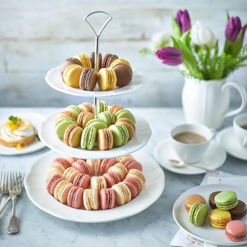 brioche pasquier mini lime,vanilla,chocolate,cream and strawberry macarons on a tiered cake tray next to a cup of tea and purple and white tulips 