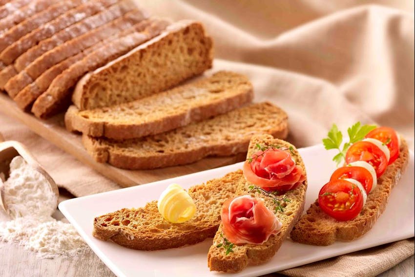 a sliced brown loaf of brioche pasquier bread with a swirl of butter, roses of parma ham and chopped vine tomatoes 