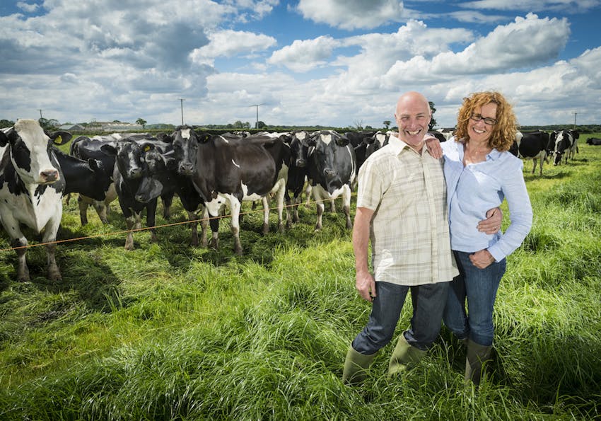 Ian and Lesley Buxton posing with arms around each other in front of a field of cows 
