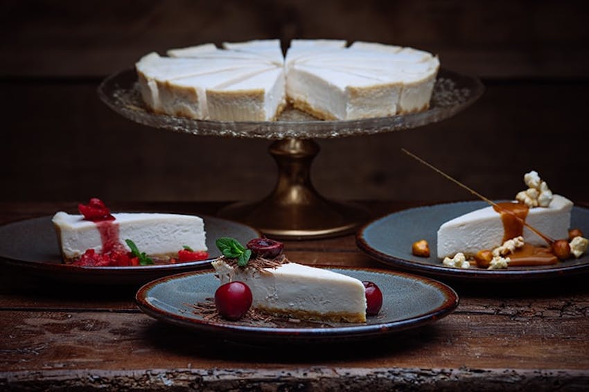 a whole central foods new york style cheesecake on a clear plated stand with three slices on grey plates topped with sauce berries and fresh mint 