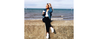 a young girl in black jeans a white top white trainers and a denim jacket on the beach wallfront holding a dairy free coffee