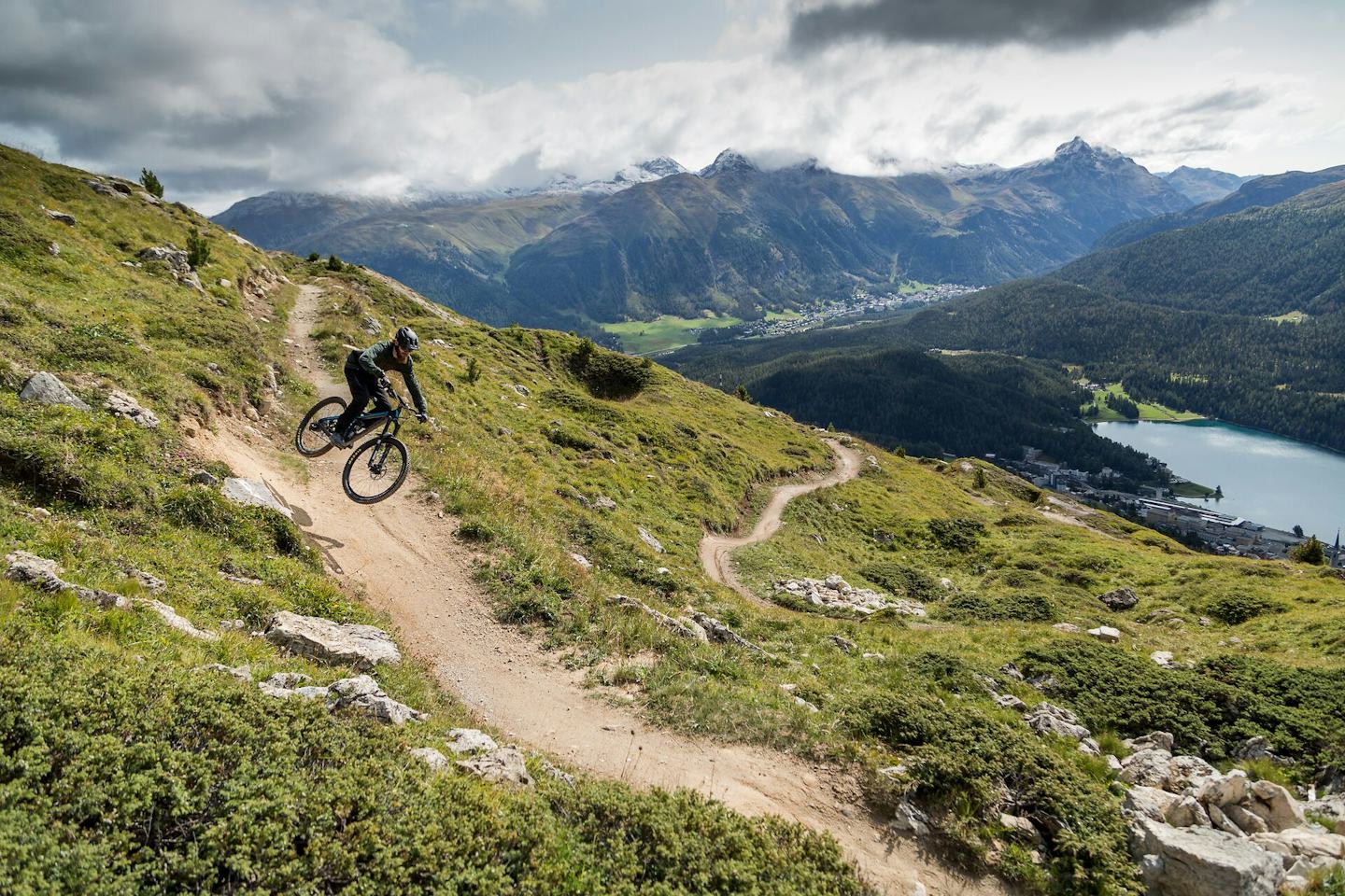 Ein Mountainbiker fährt auf einem Flowtrail ins Tal hinunter. Zu sehen sind grüne Wiesen und die Aussicht auf den St.-Moritz-See im Tal, begleitet von der spektakulären Alpenlandschaft und einem bewölkten Himmel.