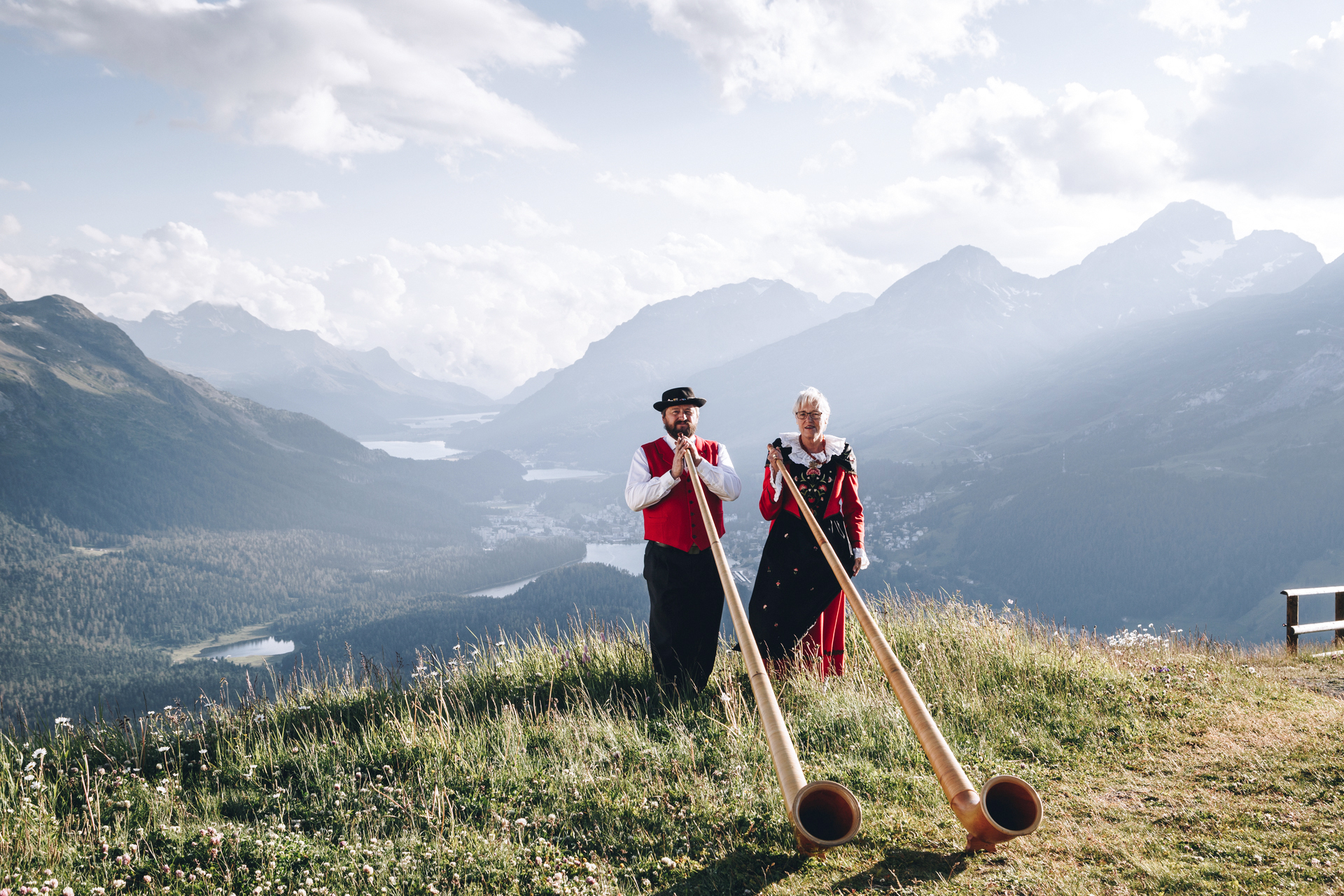 Auf Muottas Muragl stehen ein Mann und eine Frau in traditioneller Kleidung und halten jeweils ein Alphorn. Im Hintergrund liegt die weite Berglandschaft des Oberengadins mit seinen Seen und Gipfeln. Der leicht bewölkte Himmel verleiht der Szenerie eine leicht mystische Stimmung.