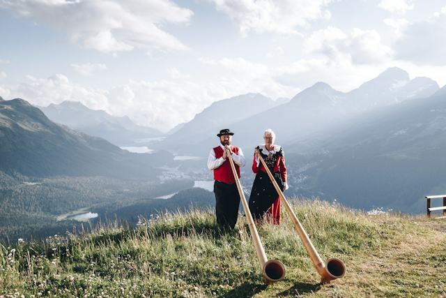 Alphorn Sunset-Apéro | Romantik Hotel Muottas Muragl Auf Muottas Muragl stehen ein Mann und eine Frau in traditioneller Kleidung und halten jeweils ein Alphorn. Im Hintergrund liegt die weite Berglandschaft des Oberengadins mit seinen Seen und Gipfeln. Der leicht bewölkte Himmel verleiht der Szenerie eine leicht mystische Stimmung.