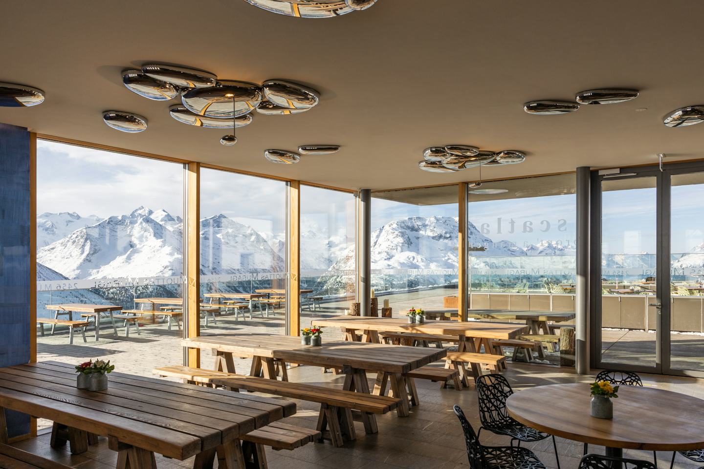 Das Restaurant Scatla – pront’ a manger auf Muottas Muragl präsentiert sich mit einem modernen, hellen Innenraum, Holztischen und einer grossen bodentiefen Fensterfront, die den Blick auf die verschneite Engadiner Bergwelt freigibt.
