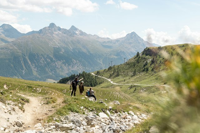 Eine Familie steht auf einem Wanderweg auf Corviglia und blickt ins Engadiner Tal. Der Vater zeigt seiner Frau und seinen zwei Kindern etwas in der Ferne. Der Hang ist mit Steinen und grünen Wiesen bedeckt, während sich auf der anderen Seite die Berge des Oberengadins erstrecken.