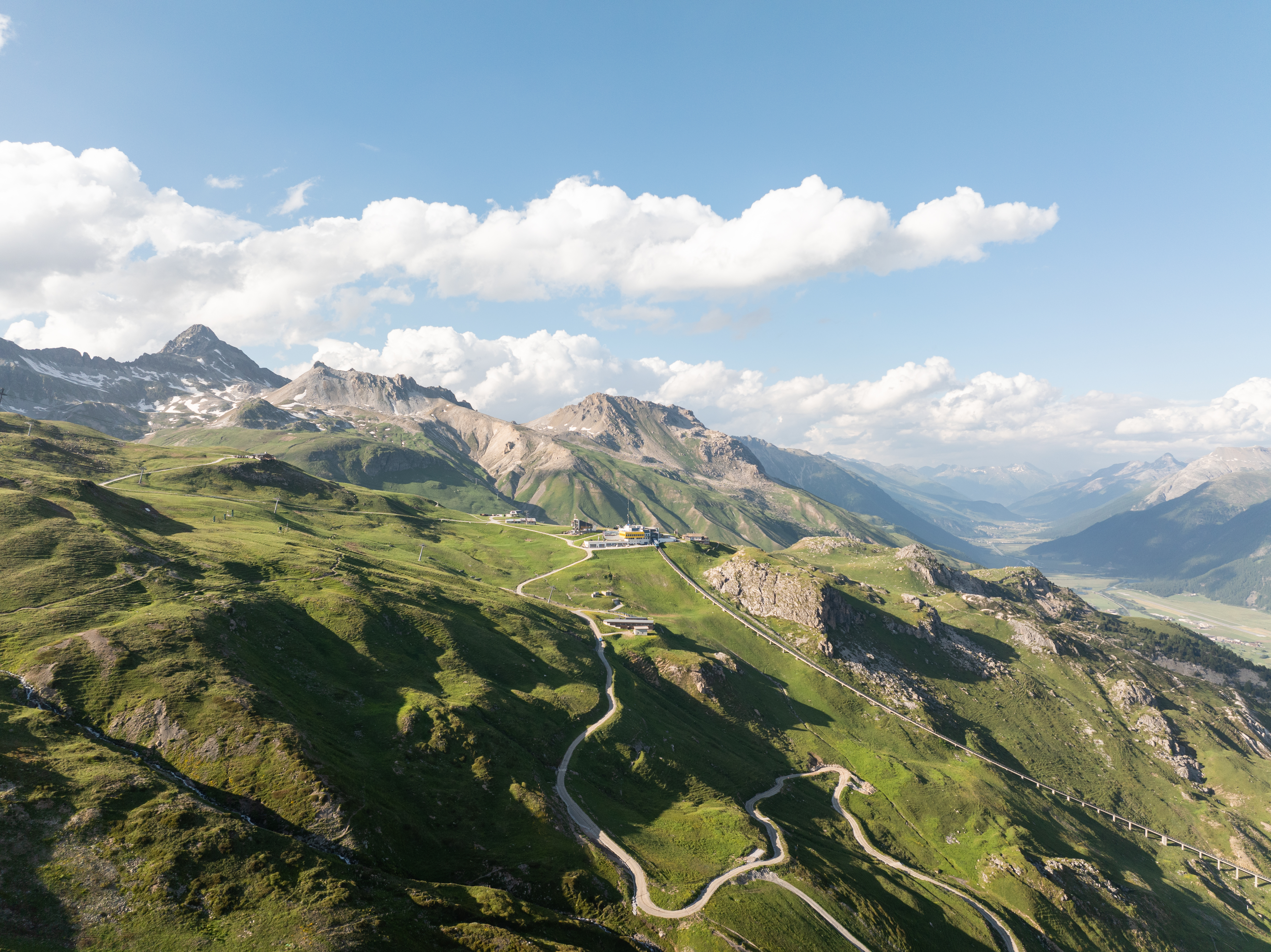 Aufnahme der Corviglia oberhalb von St. Moritz mit der markanten gelben Bergstation, grünen Alpwiesen, Serpentinenstrasse und weitem Blick über die Engadiner Berglandschaft unter blauem Himmel.