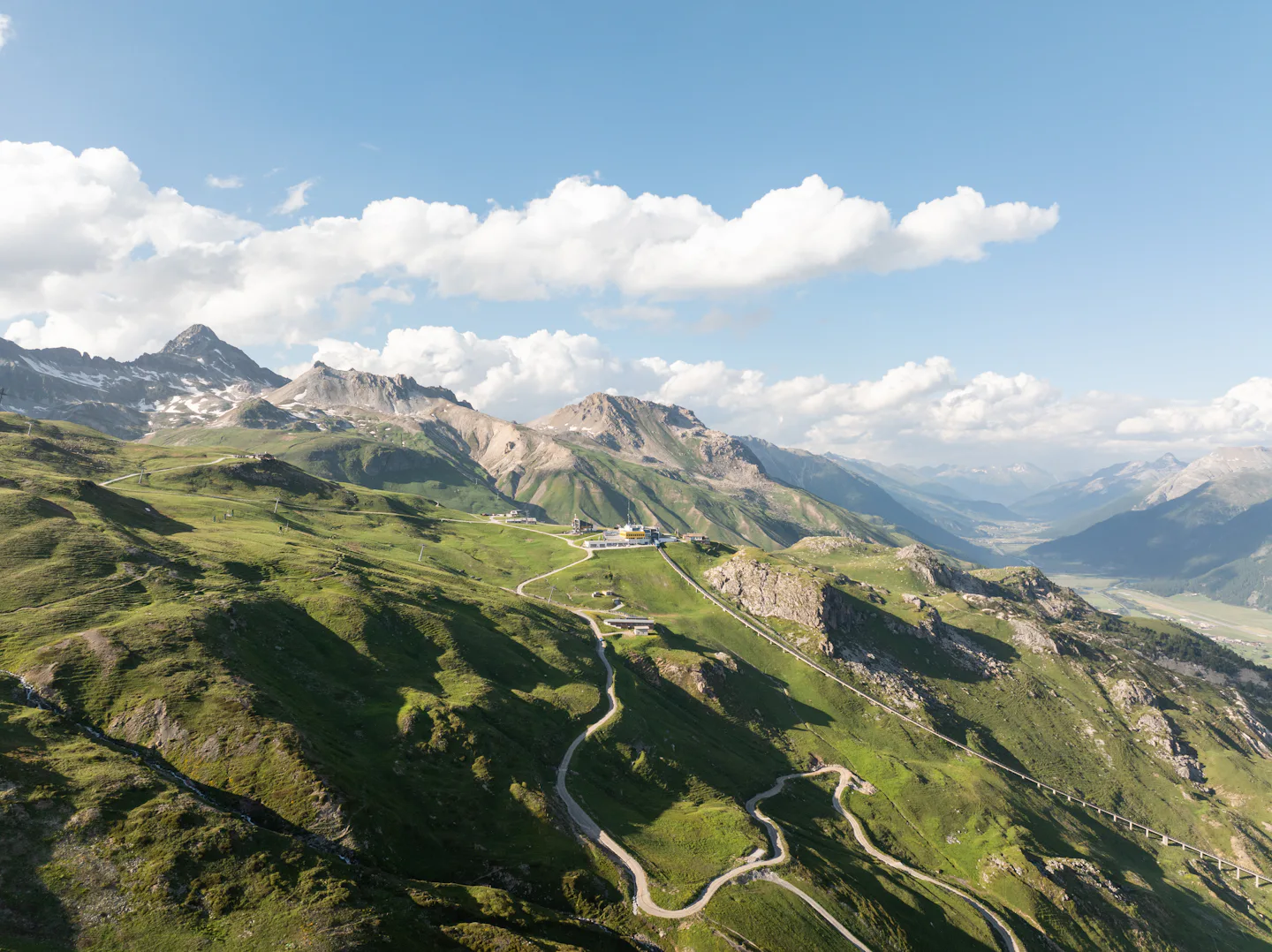 Landscape Corviglia | Sommer Aufnahme der Corviglia oberhalb von St. Moritz mit der markanten gelben Bergstation, grünen Alpwiesen, Serpentinenstrasse und weitem Blick über die Engadiner Berglandschaft unter blauem Himmel.