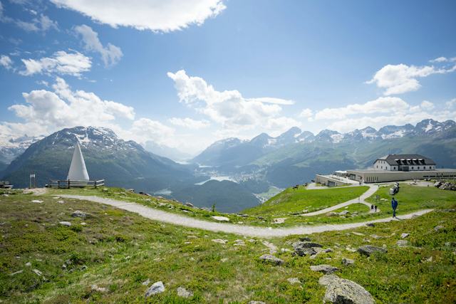 Die Berglandschaft des Engadins, gesehen von Muottas Muragl. Im Vordergrund liegt das Romantik Hotel Muottas Muragl. Dahinter grüne Hänge, Täler mit den Oberengadiner Seen und die Gipfel der Schweizer Alpen unter klarem Himmel. Links im Bild ist ein Tropfen aus Naturstein, Mörtel und weissem Marmor als Kunstwerk auf dem Kunstpfad "Senda d’Inspiraziun" zu sehen.