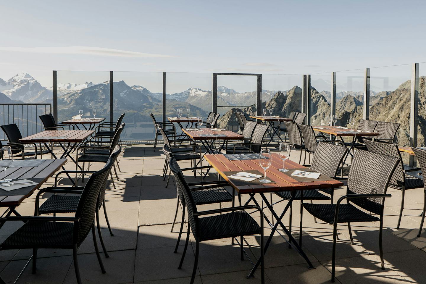 Die Panoramaterrasse des Bergrestaurants Piz Nair 10000 Feet auf 3057 Metern. In der Sonne stehen gedeckte Tische mit Blick auf die Alpenlandschaft rund um den St. Moritzer Hausberg Piz Nair. Im Hintergrund sind Berge mit weissen Gipfeln und ein weiter blauer und wolkenloser Himmel zu sehen.