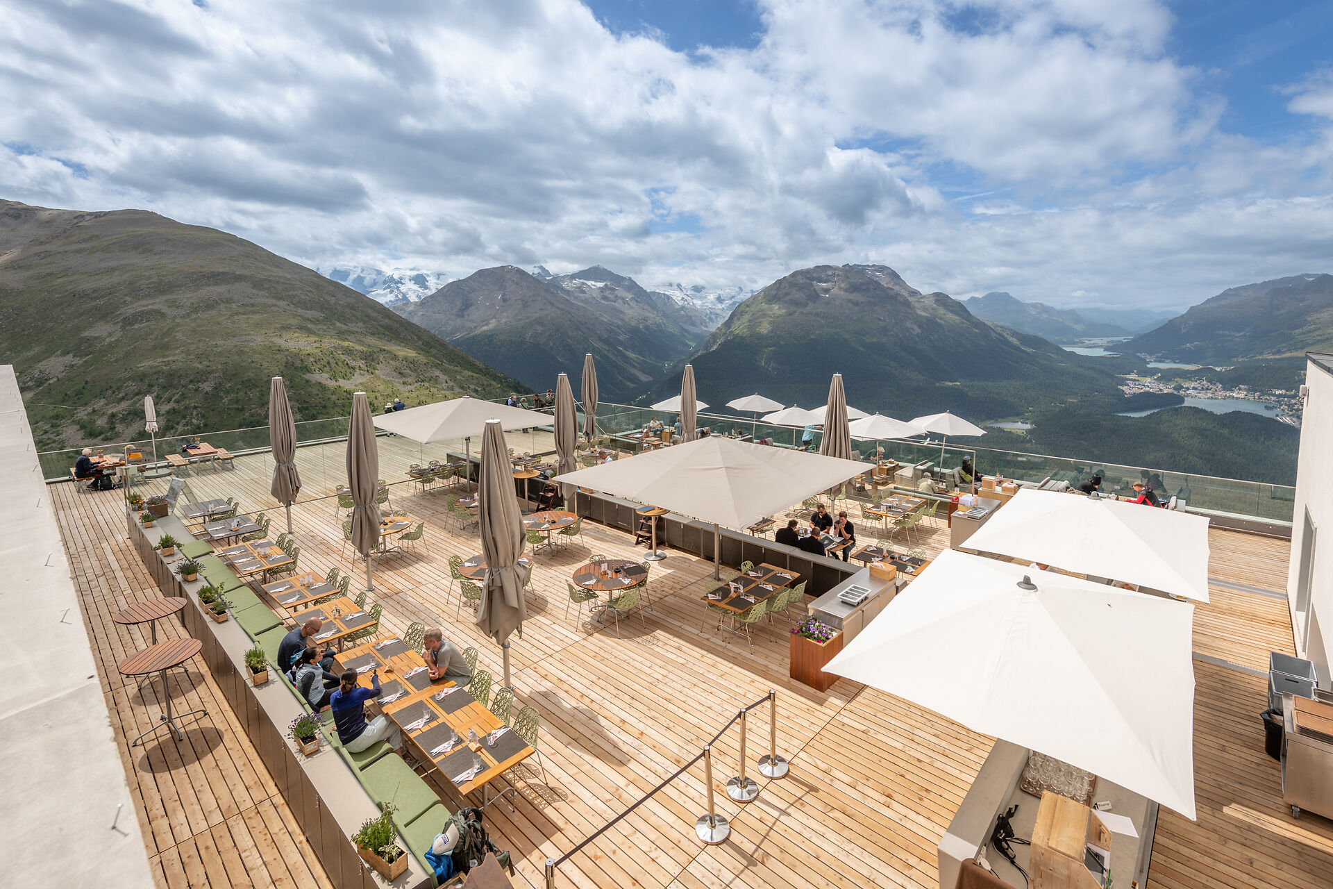 Die grosszügige Panoramaterrasse des Romantik Hotels Muottas Muragl bietet zahlreiche Sitzplätze im Freien. Sie ist mit Tischen, Stühlen und Sonnenschirmen ausgestattet und liegt an einem Hang. Im Hintergrund erstreckt sich die weitläufige alpine Berglandschaft des Engadins mit Tälern und Gipfeln unter klarem Himmel.