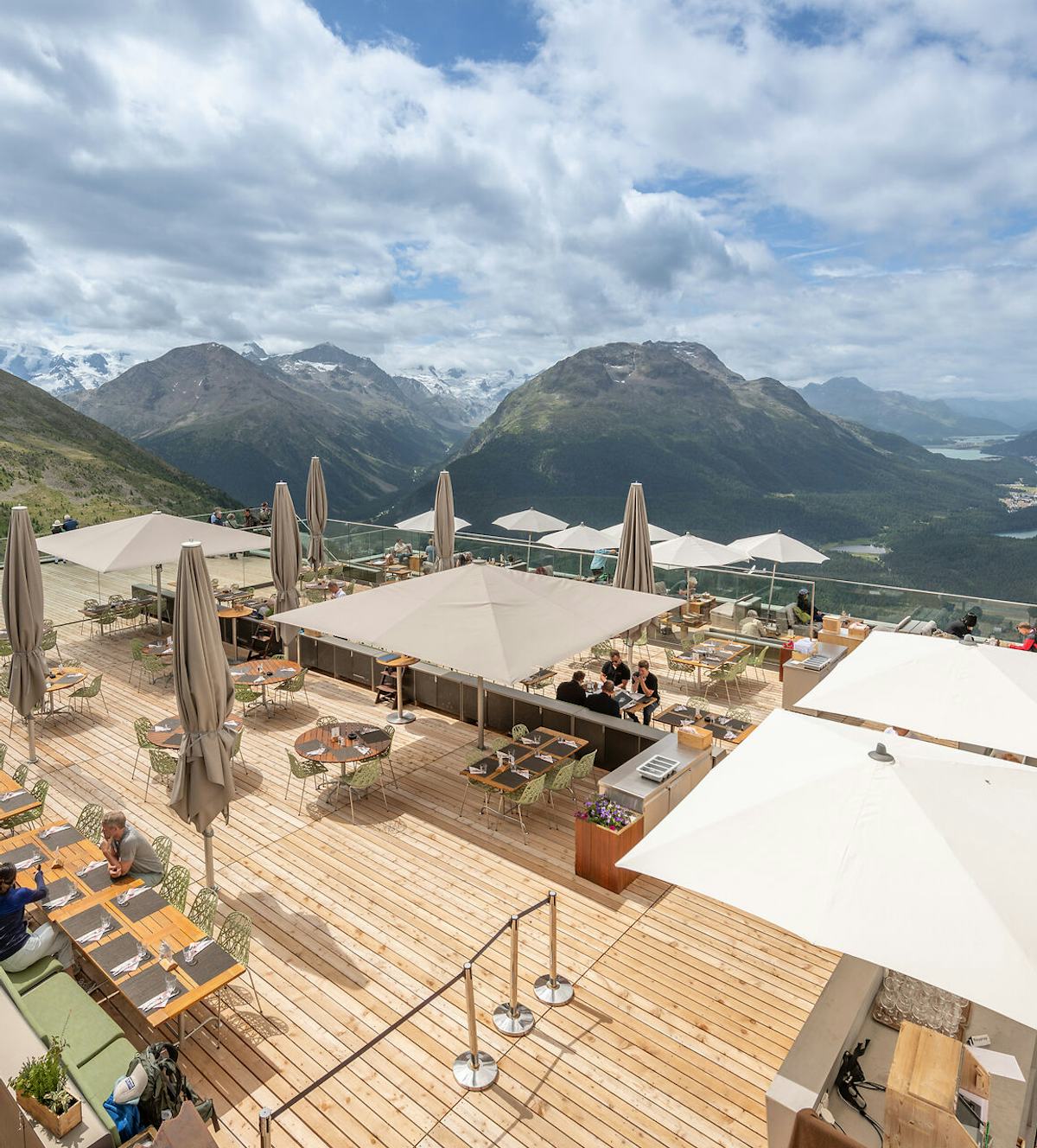 Panoramaterrasse | Romantik Hotel Muottas Muragl | Sommer Die grosszügige Panoramaterrasse des Romantik Hotels Muottas Muragl bietet zahlreiche Sitzplätze im Freien. Sie ist mit Tischen, Stühlen und Sonnenschirmen ausgestattet und liegt an einem Hang. Im Hintergrund erstreckt sich die weitläufige alpine Berglandschaft des Engadins mit Tälern und Gipfeln unter klarem Himmel.