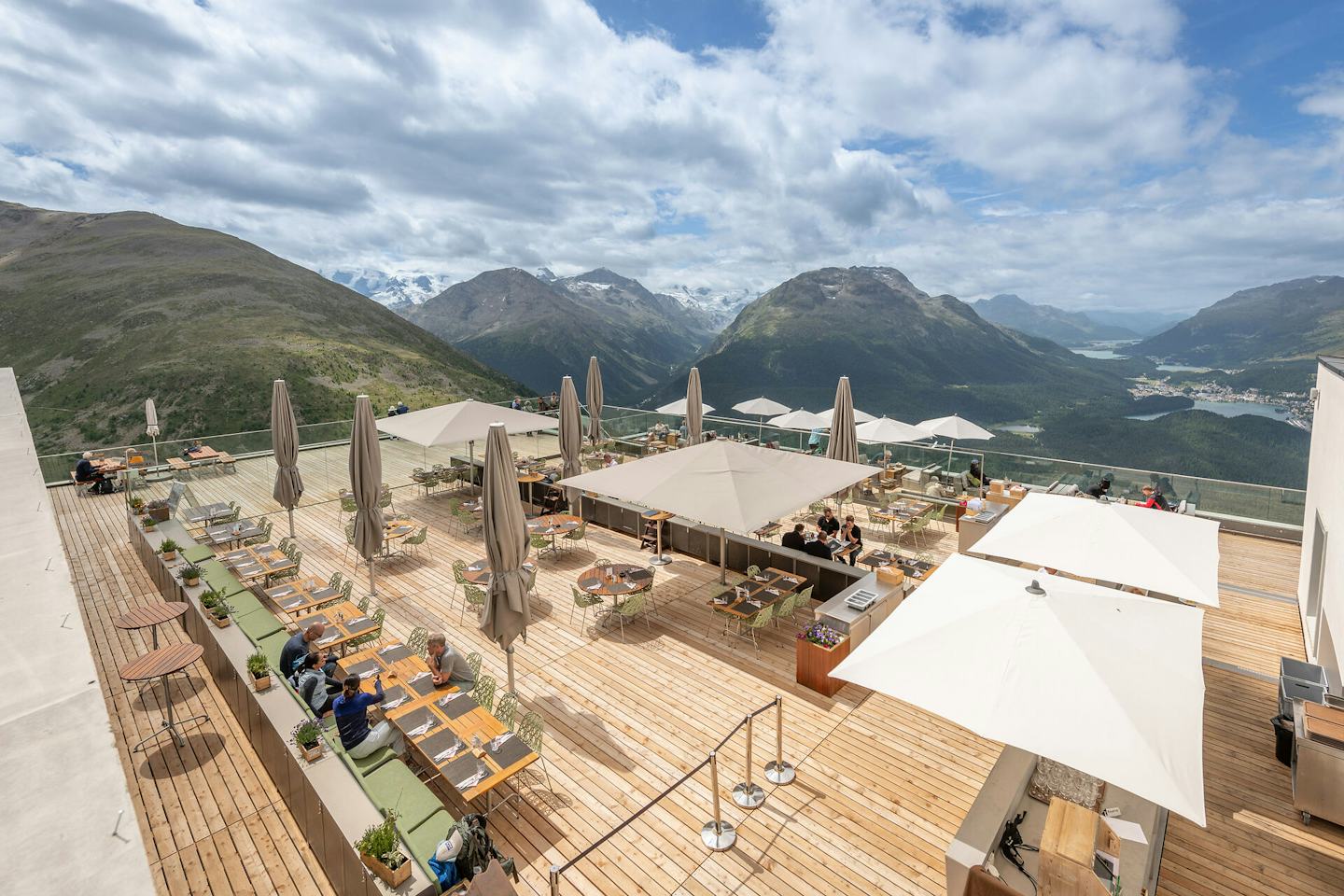 Die grosszügige Panoramaterrasse des Romantik Hotels Muottas Muragl bietet zahlreiche Sitzplätze im Freien. Sie ist mit Tischen, Stühlen und Sonnenschirmen ausgestattet und liegt an einem Hang. Im Hintergrund erstreckt sich eine weitläufige alpine Berglandschaft mit Tälern und Gipfeln unter klarem Himmel.