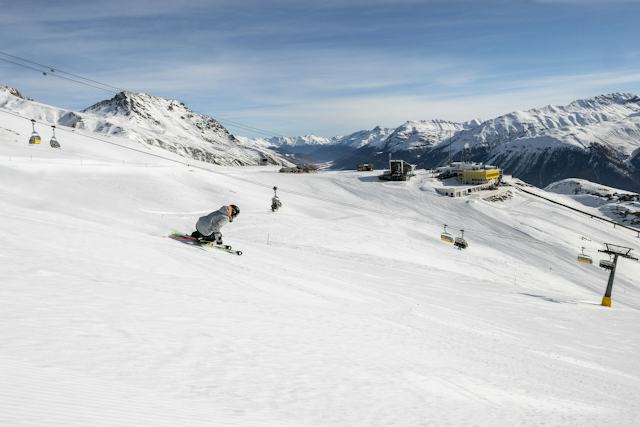 Skifahren Corviglia Ein einzelner Skifahrer carvt auf perfekt präparierten Pisten an der Bergstation Corviglia vorbei. Im Bild sind neben ihm eine Sesselbahn und im Hintergrund die schneebedeckten Gipfel zu sehen.